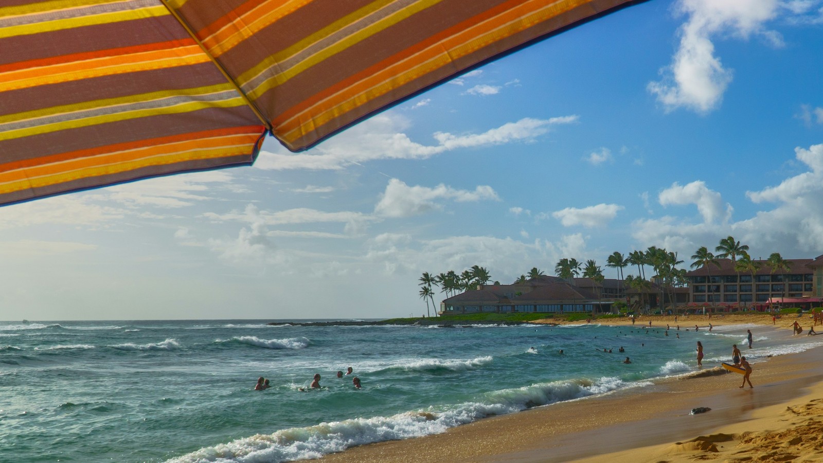 Beachgoers and waves at Poʻipū Beach, with a resort and palm trees along the shore.